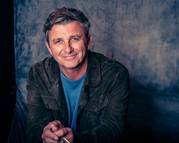 A smiling man with gray-streaked hair sits in front of a neutral background. He wears a jacket and a T-shirt and looks friendly at the camera.