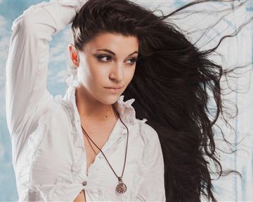 A young woman with long, flowing hair is wearing a white shirt. She looks thoughtful and has a necklace around her neck.