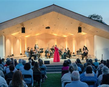 A musician in a red dress stands at the center of a stage and sings. The audience sits eagerly in front of her, surrounded by a beautiful evening backdrop.