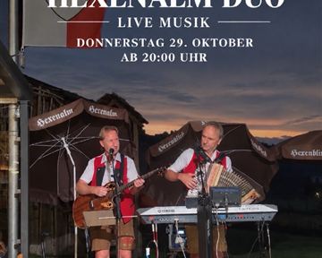 Two musicians are performing live on an outdoor stage. In the background, a beautiful sunset and the mountains can be seen.