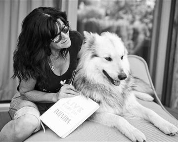 Eine Frau sitzt entspannt auf einer Liege und streichelt ihren Hund. Sie hält ein Notizbuch in der Hand und lächelt zufrieden.
