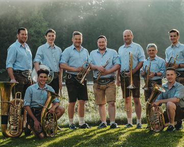 A group of musicians is standing outdoors and wearing traditional costumes. They are holding various wind instruments and smiling at the camera.