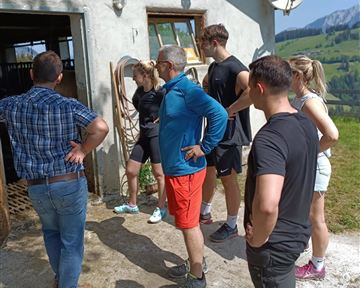 A group of people is standing outside in front of a farmhouse. They are looking at something curiously and seem to be interested.