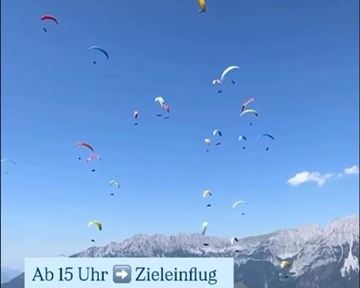 Ein klarer blauer Himmel mit vielen bunten Gleitschirmfliegern. Im Hintergrund sind Berge und grüne Wiesen zu sehen.