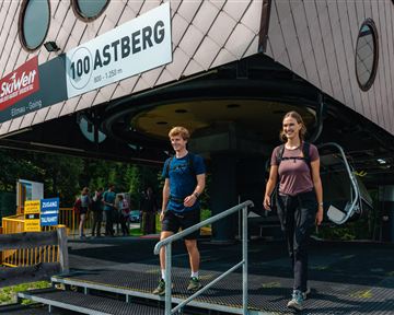 Zwei Wanderer verlassen eine moderne Seilbahnstation. Im Hintergrund sind weitere Personen zu sehen, die sich auf den Weg machen.
