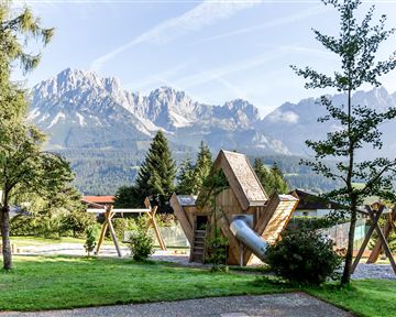 Ein schöner Spielplatz in einem Park mit einem einzigartigen Spielgerät und schattenspendenden Bäumen. Im Hintergrund sind beeindruckende Berge zu sehen.