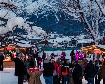 A wintry Christmas market with people in warm clothes. In the background, snow-covered mountains can be seen.