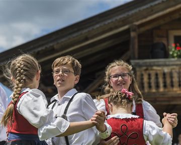 Kinder in traditioneller Tracht tanzen zusammen vor einem rustikalen Holzhaus. Der Himmel ist blau und die Stimmung ist fröhlich.