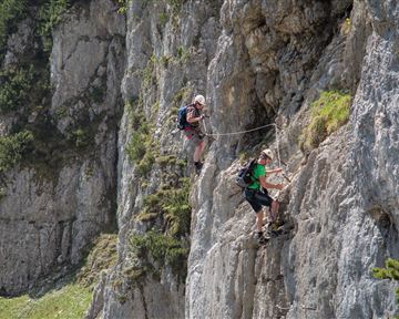 Two climbers rappelling down a rock face. The surroundings are green and mountainous, and the sun is shining.