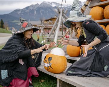 Zwei Frauen in Hexenkostümen stehen vor einem Holzregal mit Kürbissen. Sie scheinen einen Kürbis mit einem Gesicht zu betrachten und wirken dabei begeistert.