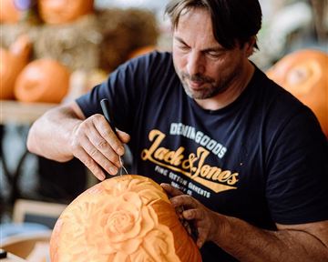 A man is carving a pumpkin with a tool. In the background, more pumpkins and straw bales are visible.