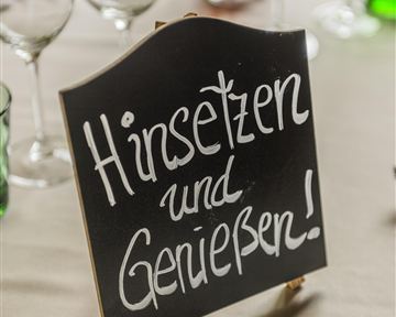 Ein schwarzes Schild mit der Aufschrift "Hinsetzen und Genießen!" steht auf einem Tisch. Um das Schild sind Gläser in verschiedenen Farben angeordnet.