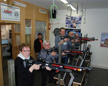 A group of five people stands in a room and conducts training sessions with sports rifles. Offices and posters are visible in the background.