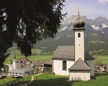 Eine malerische Kirche mit einer charakteristischen Kuppel steht in einer grünen Landschaft. Im Hintergrund sind majestätische Berge zu sehen, die die idyllische Szene umrahmen.