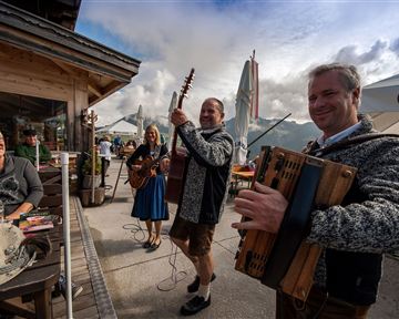 Eine fröhliche Musikkapelle unterhalb eines Bergs spielt traditionelle Lieder. Gäste genießen die Atmosphäre in einem gemütlichen Außenbereich.