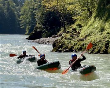 Drei Kajakfahrer paddeln auf einem ruhigen Fluss. Im Hintergrund sind grüne Bäume und eine sonnige Landschaft zu sehen.