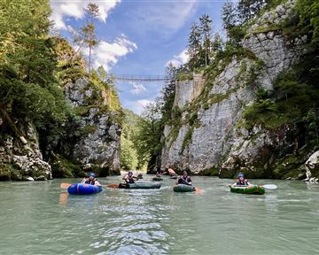 A group of people is swimming on rafts in a clear river, surrounded by tall rocks and trees. In the background, a pedestrian bridge is visible.