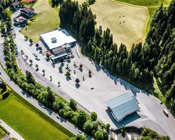 Ein weites Parkplatzareal mit mehreren Autos und einer kleinen Verkaufsstelle. Umgeben von grünen Wäldern und sanften Hügeln.