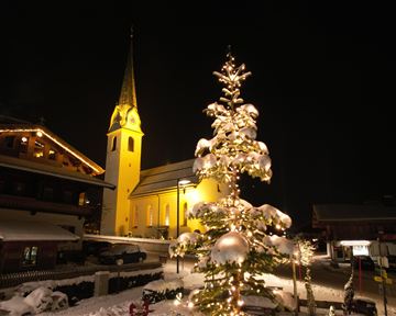 Ein festlich geschmückter Weihnachtsbaum steht im Schnee, beleuchtet von warmem Licht. Im Hintergrund ist eine Kirche mit einem hohen Turm zu sehen, die ebenfalls in warmes Licht getaucht ist.