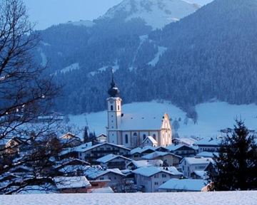 Eine malerische Winterlandschaft mit einem Dorf im Schnee. Am Horizont erhebt sich ein majestätischer Berg und eine Kirche prägt die Szene.