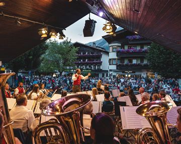 An open-air concert with an orchestra on a stage. Many spectators enjoy the performance outdoors.