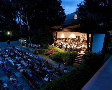 Ein Konzert im Freien mit einem Orchester auf einer Bühne. Viele Zuschauer sitzen auf Bänken und genießen die Musik in einer gemütlichen Abendstimmung.
