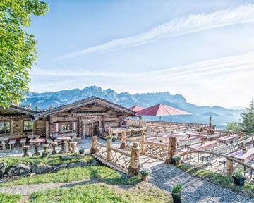 Ein rustikales Bergrestaurant mit Holzkonstruktion und schöner Außenterrasse. Im Hintergrund sind majestätische Berge und ein klarer Himmel zu sehen.