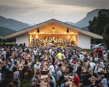 A large crowd is enjoying an outdoor concert. A music band is playing on stage, while in the foreground there are many tables and chairs.