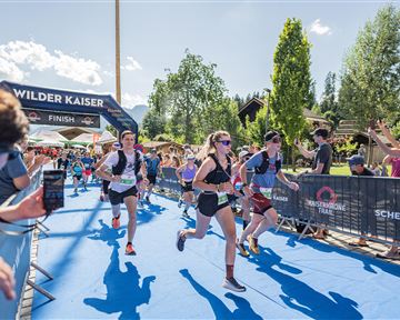 Eine Gruppe von Läufern erreicht das Ziel bei einem Wettlauf. Zuschauer stehen an der Strecke und feuern die Teilnehmer an.