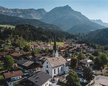 Eine malerische Dorfansicht mit einer weißen Kirche im Vordergrund und den Alpen im Hintergrund. Die Umgebung ist grün und bergig, ideal für Naturliebhaber.