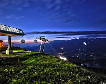 Eine Bergbahn mit Gondeln bei Nacht in der Natur. Im Hintergrund sind die silhouettierenden Berge und der klare Nachthimmel zu sehen.
