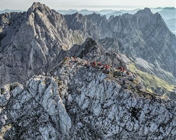 Ein atemberaubender Blick auf einen zerklüfteten Gebirgskamm mit vielen Menschen auf dem Gipfel. Die Landschaft ist von majestätischen Bergen und einer klaren Himmelspanne umgeben.