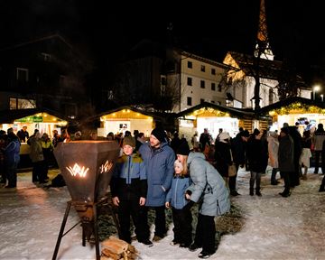 Ein winterlicher Marktplatz bei Nacht mit festlicher Beleuchtung. Menschen genießen die Atmosphäre und die Stände im Hintergrund.