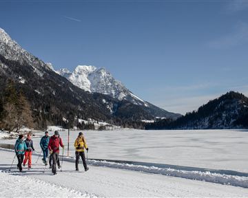 A group of five people is hiking along the shore of a frozen lake. In the background, snow-covered mountains and clear blue skies are visible.