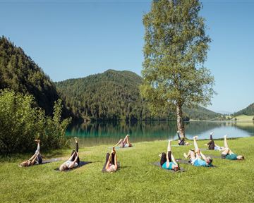 A group of people is doing stretching exercises on a green meadow next to a clear lake. In the background, wooded hills and a blue sky can be seen.