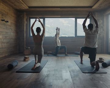 Eine ruhige Yogastunde in einem Holzraum mit Blick auf die Berge. Drei Personen praktizieren verschiedene Yoga-Posen auf Matten.