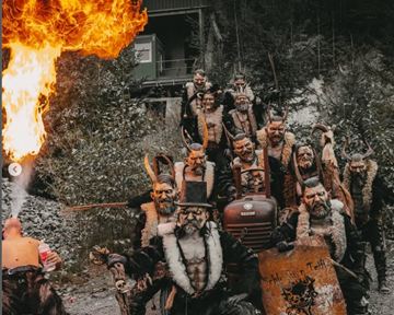 A group of masked people in eerie costumes stands outdoors. One of the participants breathes fire while the others take grim poses.