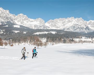 Two people are hiking through a snow-covered landscape. Impressive mountains can be seen in the background under a clear blue sky.
