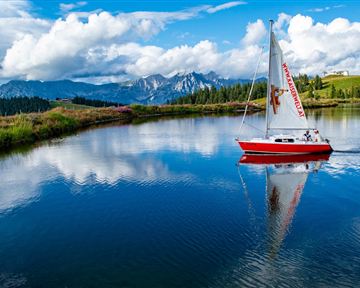 Ein rotes Segelboot schwimmt auf einem ruhigen See. Im Hintergrund sind Berge und eine schöne Wolkenlandschaft zu sehen.