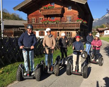 A group of six people is riding Segways in a rural setting. In the background, a traditional wooden main house with flowers can be seen.