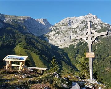 Eine schöne Berglandschaft mit hohen Gipfeln und grünem Wald. Im Vordergrund steht ein Holzkreuz und eine Informationstafel.