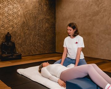 A person is performing a relaxation treatment while another person lies on a couch. The room is designed to be calming, with a Buddha and geometric wall decoration.
