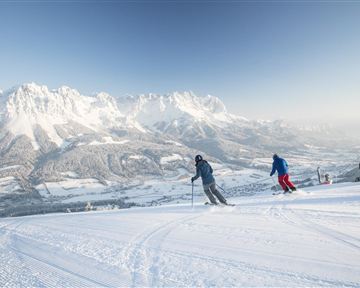 Zwei Skifahrer fahren die verschneite Piste hinunter. Im Hintergrund sind majestätische Berge und ein klarer Himmel zu sehen.