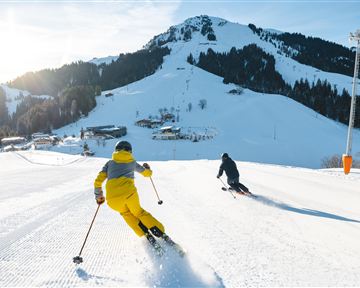 Zwei Skifahrer fahren die Piste hinunter, umgeben von schneebedeckten Bergen. Die Sonne scheint hell am blauen Himmel.