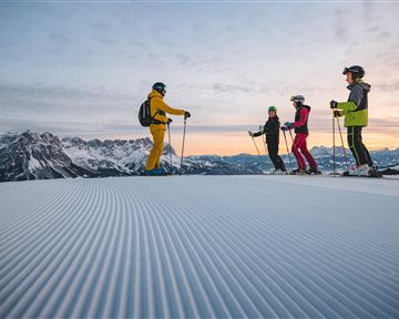 Eine Gruppe von Skifahrern steht auf einer verschneiten Piste mit Bergen im Hintergrund. Der Himmel ist klar und zeigt schöne Farben beim Sonnenuntergang.