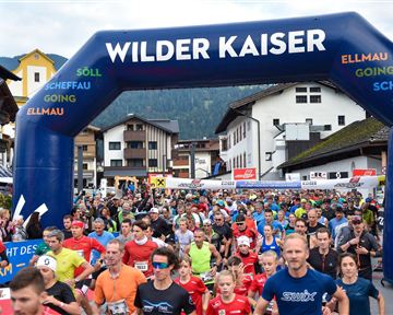 Ein großes Teilnehmerfeld beim Start eines Rennens unter einem blauen Tor mit der Aufschrift "Wilder Kaiser". Viele Läufer in bunten Shirts bewegen sich in Richtung des Starts in einer malerischen Umgebung.