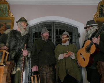 A group of four men in historical costumes stands in front of a wrought-iron gate. One of the men holds a guitar and the others have various accessories in their hands.