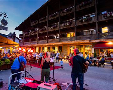 Eine belebte Straßenszene mit Musikern, die vor einem Holzgebäude auftreten. Viele Menschen sitzen draußen und genießen die Atmosphäre am Abend.