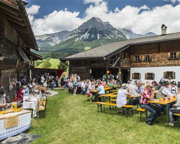 A lively mountain festival with many people sitting at tables outdoors. In the background, picturesque mountains and traditional wooden houses can be seen.