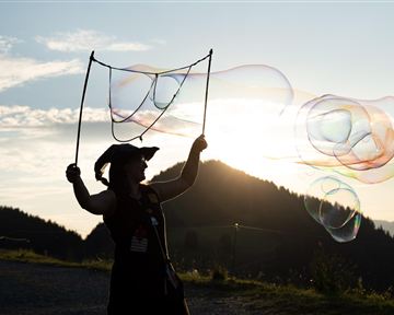 Eine Person steht im Sonnenuntergang und bläst Seifenblasen. Die Berge und der Himmel sind im Hintergrund gut sichtbar.